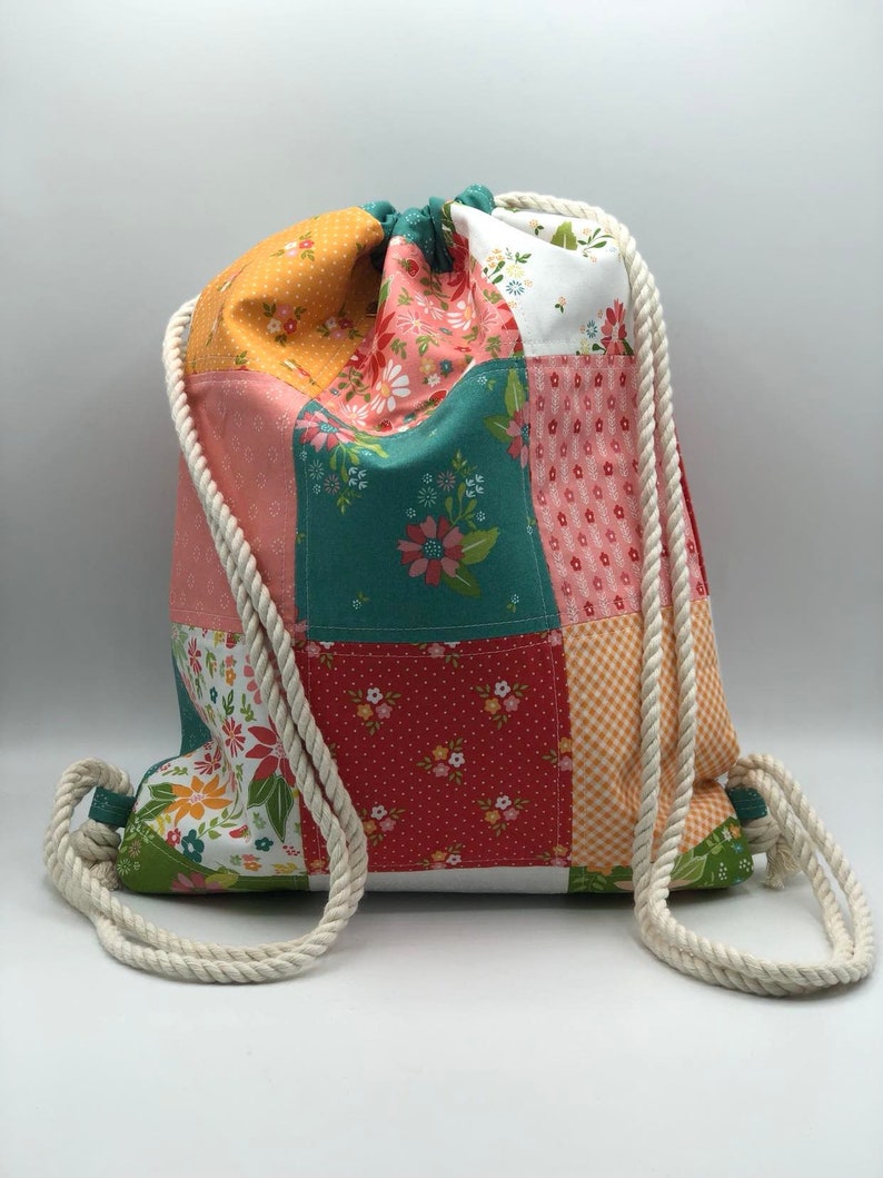 K&ouml;nnte beinhalten: Eine Patchwork-Kordeltasche mit einem Blumenmuster in den Farben Rosa, Rot, Orange, Gr&uuml;n und Wei&szlig;. Die Tasche hat einen wei&szlig;en Baumwollkordelzug.