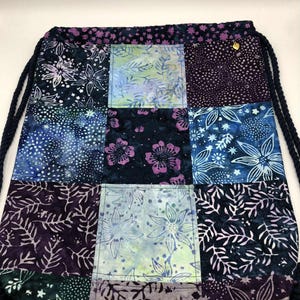May include: A patchwork drawstring bag with a variety of floral and abstract patterns in shades of blue, purple, and green. The bag features a dark blue drawstring closure and a small gold-colored accent. The bag is made of fabric.