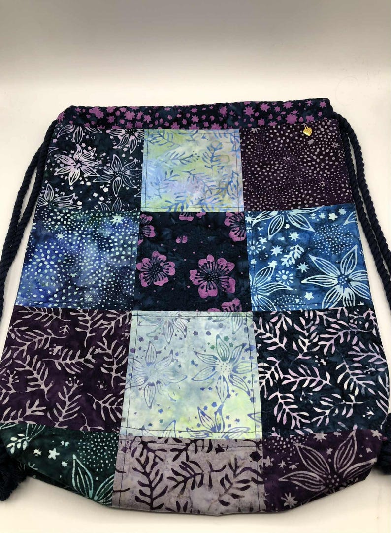 May include: A patchwork drawstring bag with a variety of floral and abstract patterns in shades of blue, purple, and green. The bag features a dark blue drawstring closure and a small gold-colored accent. The bag is made of fabric.