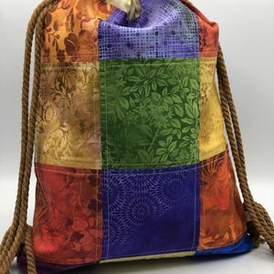 May include: A colorful drawstring backpack with a patchwork design. The bag features squares of fabric in various colors, including red, orange, green, and purple, with floral and patterned designs. The drawstring is made of a thick, brown rope.
