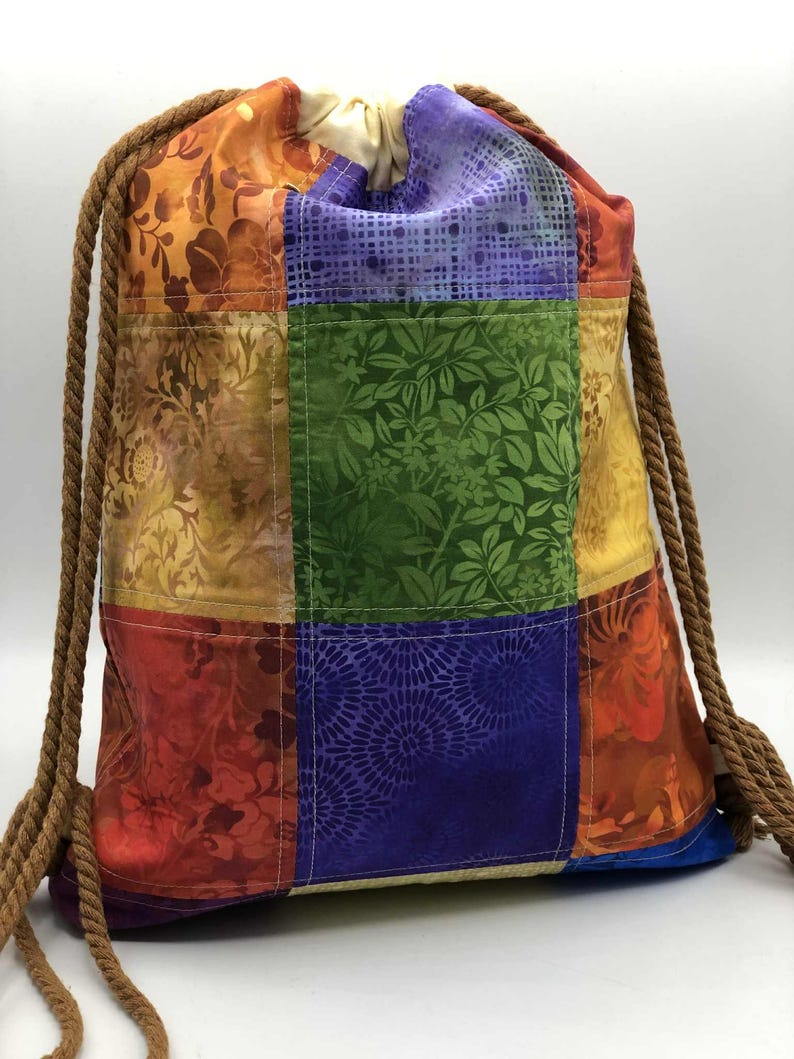 以下が含まれることがあります： パッチワークデザインのカラフルな巾着バックパック。赤、オレンジ、緑、紫など、さまざまな色の生地の正方形が特徴で、花柄や模様のデザインが施されています。巾着は太い茶色のロープでできています。