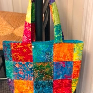 May include: A colorful patchwork tote bag with a variety of vibrant fabrics. The bag features a mix of red, blue, orange, and green squares, with matching handles. The bag is designed for carrying items.