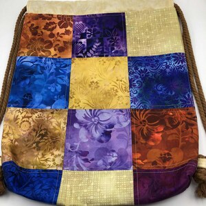 以下が含まれることがあります： オレンジ、青、紫、金色の様々な花柄のパッチワーク巾着袋。バッグはベージュのトップと茶色のロープの巾着が付いています。バッグは布製です。