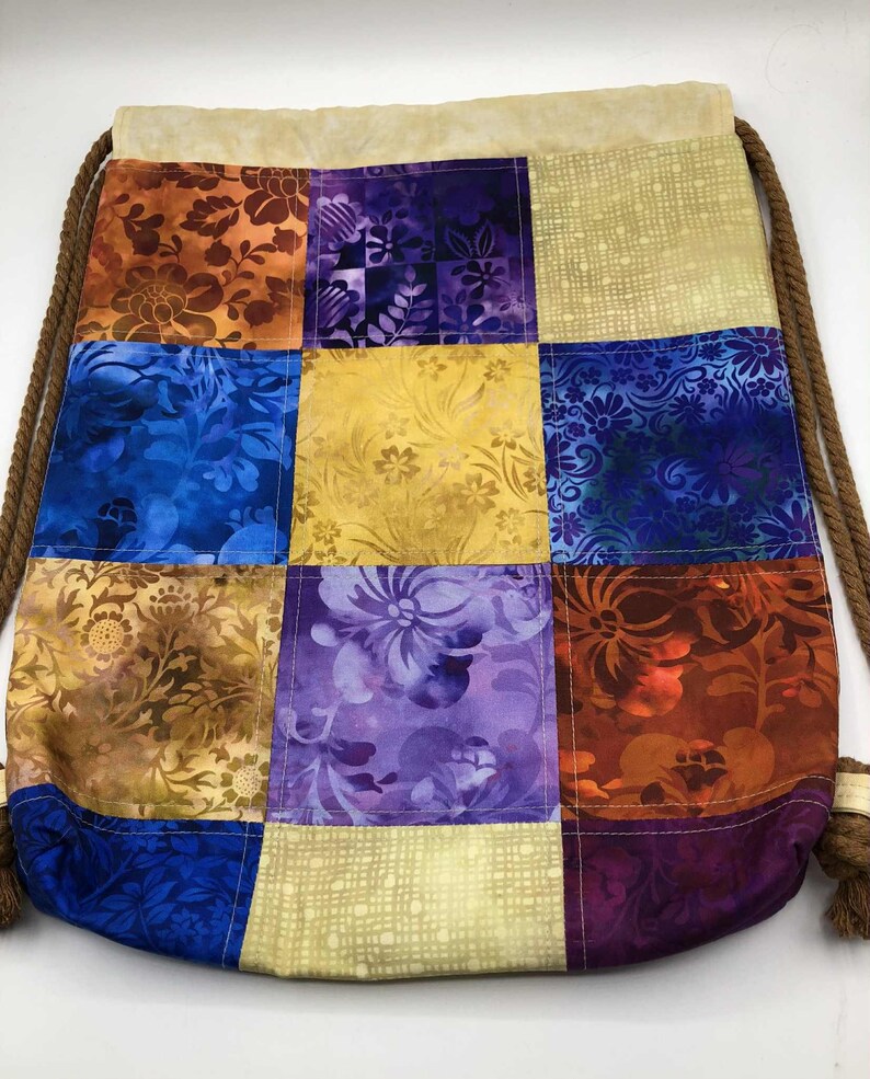 以下が含まれることがあります： オレンジ、青、紫、金色の様々な花柄のパッチワーク巾着袋。バッグはベージュのトップと茶色のロープの巾着が付いています。バッグは布製です。