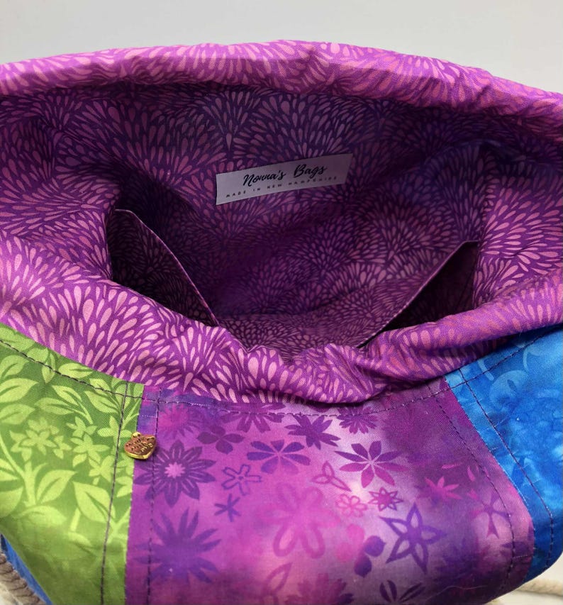 May include: A colourful tote bag with a drawstring closure. The bag has a patchwork design with green, purple, and blue floral patterns. The interior is lined with purple fabric. The bag has a label that reads "Nova's Bags".