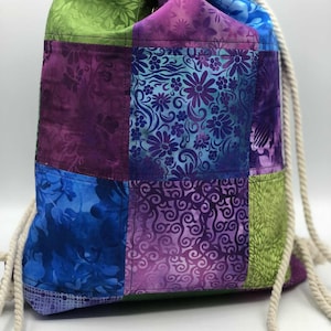 May include: A patchwork drawstring bag in shades of blue, purple, and green. The bag features floral and abstract patterns. The drawstring is made of a thick, off-white rope. The bag is designed for carrying items.