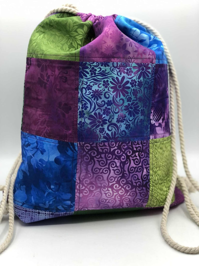 May include: A patchwork drawstring bag in shades of blue, purple, and green. The bag features floral and abstract patterns. The drawstring is made of a thick, off-white rope. The bag is designed for carrying items.