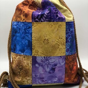 以下が含まれることがあります： 青、紫、金、オレンジの色合いの様々な花柄が特徴の、カラフルなパッチワーク巾着バックパック。茶色のロープのドローストリングが付いており、生地で作られています。