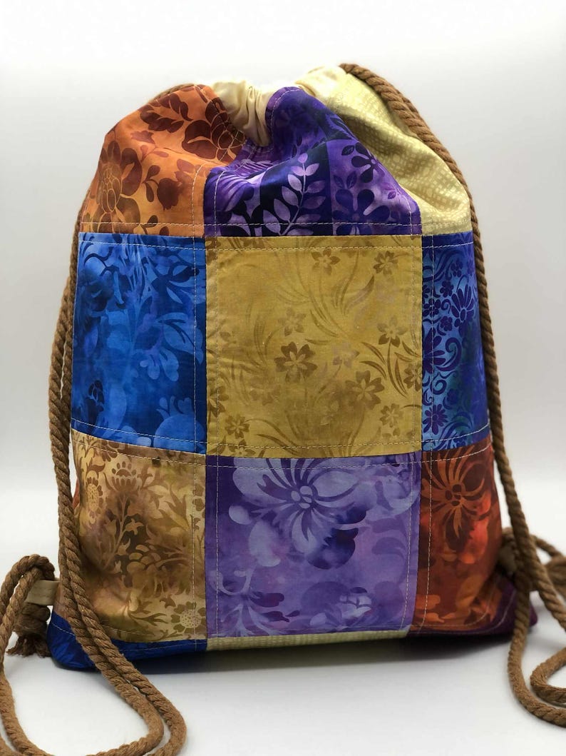 以下が含まれることがあります： 青、紫、金、オレンジの色合いの様々な花柄が特徴の、カラフルなパッチワーク巾着バックパック。茶色のロープのドローストリングが付いており、生地で作られています。