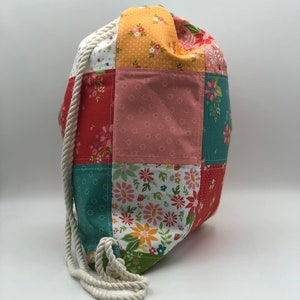 K&ouml;nnte beinhalten: Eine Patchwork-Kordeltasche mit einem wei&szlig;en Seilgriff. Die Tasche besteht aus verschiedenen Stoffen mit Blumenmustern in Rot-, Rosa-, Gelb-, Gr&uuml;n- und Wei&szlig;t&ouml;nen.