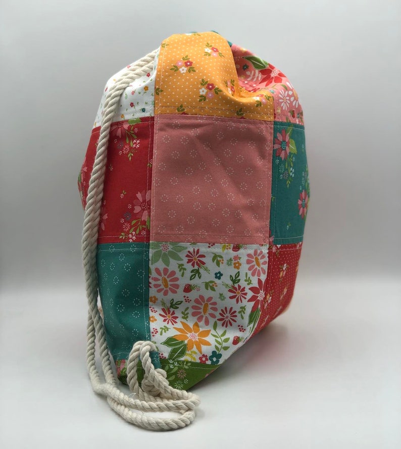 K&ouml;nnte beinhalten: Eine Patchwork-Kordeltasche mit einem wei&szlig;en Seilgriff. Die Tasche besteht aus verschiedenen Stoffen mit Blumenmustern in Rot-, Rosa-, Gelb-, Gr&uuml;n- und Wei&szlig;t&ouml;nen.