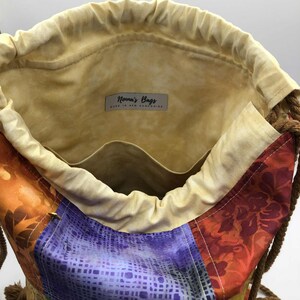 以下が含まれることがあります： パッチワークデザインのカラフルな巾着袋。オレンジ、紫、緑、黄色の様々な生地を使用しています。内側は薄いタン色で、紐で開閉し、ロープの持ち手が付いています。「Nonna's Bags」と書かれたラベルが付いています。