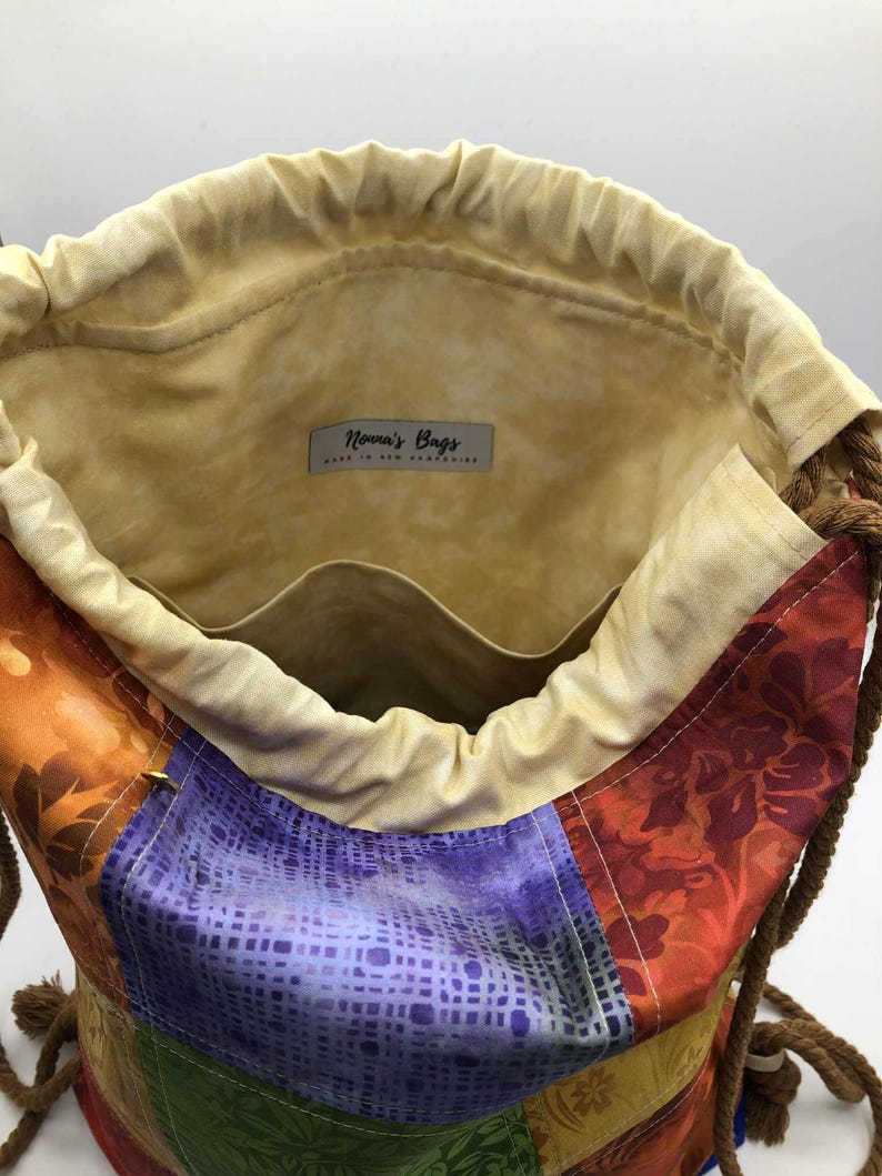 以下が含まれることがあります： パッチワークデザインのカラフルな巾着袋。オレンジ、紫、緑、黄色の様々な生地を使用しています。内側は薄いタン色で、紐で開閉し、ロープの持ち手が付いています。「Nonna's Bags」と書かれたラベルが付いています。