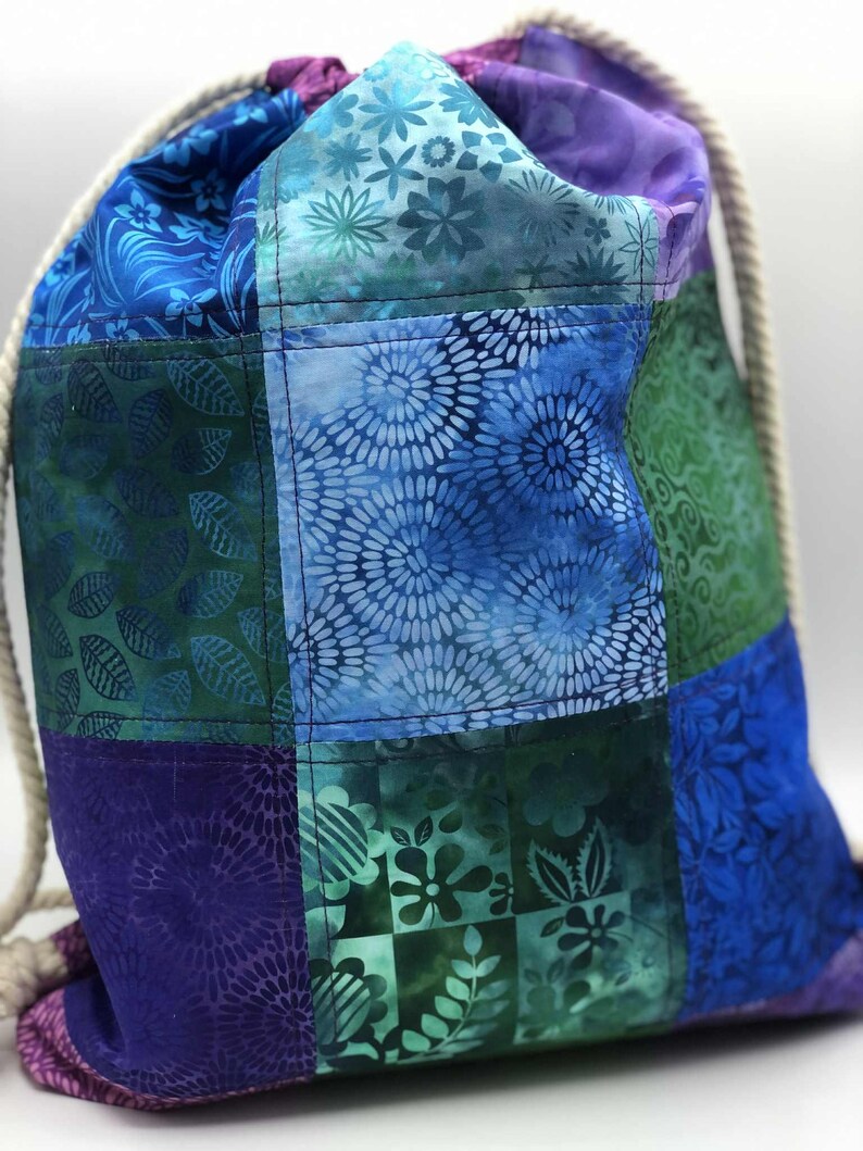 May include: A patchwork drawstring bag in shades of blue, green, and purple. The bag features floral and leaf patterns. A thick, white rope serves as the drawstring and shoulder straps. The bag is made of fabric.