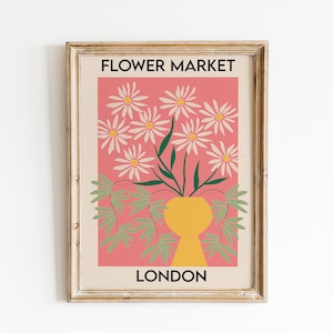 May include: Framed art print featuring a floral design. The artwork displays white daisies in a yellow vase against a pink background. The words "FLOWER MARKET" and "LONDON" are printed in black. The frame is a light brown color.