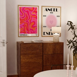 May include: Two framed posters on a wall above a wooden dresser. The poster on the left has a pink and orange abstract swirl design with the word "Euphoria" at the bottom. The poster on the right has a pink and white background with the word "Angel" at the top and the words "Soul Energy" at the bottom. There are numbers around a pink heart in the center of the poster.