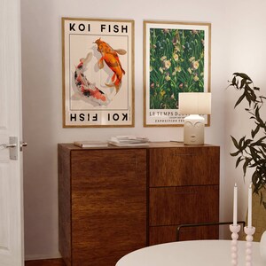 Op de afbeelding: Twee ingelijste posters op een witte muur, een met een kleurrijke illustratie van twee koi-vissen en de tekst "KOI FISH", de andere met een groene bloemenillustratie en de tekst "LE TEMPS D'AUTUMN - EXPOSITION TEA ROOM". Onder de posters staat een houten kast met drie lades. Op de kast staat een witte keramische lamp met een gezichtsontwerp. Op de voorgrond staat een witte tafel met een roze kaarsenhouder.