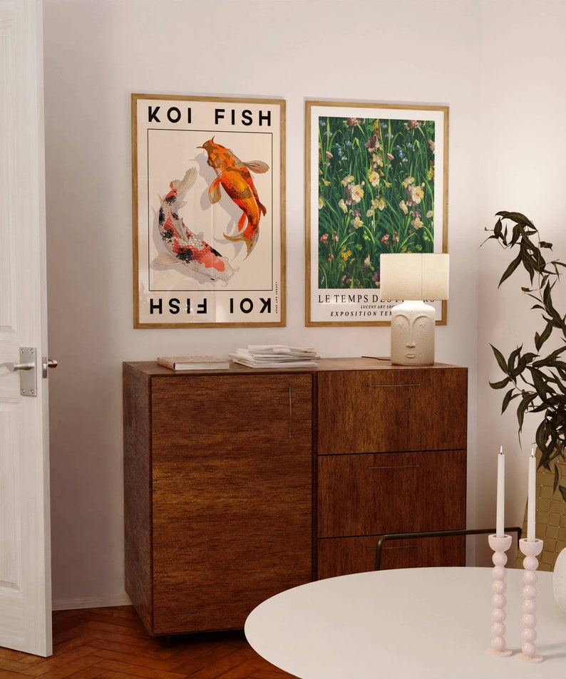 Op de afbeelding: Twee ingelijste posters op een witte muur, een met een kleurrijke illustratie van twee koi-vissen en de tekst "KOI FISH", de andere met een groene bloemenillustratie en de tekst "LE TEMPS D'AUTUMN - EXPOSITION TEA ROOM". Onder de posters staat een houten kast met drie lades. Op de kast staat een witte keramische lamp met een gezichtsontwerp. Op de voorgrond staat een witte tafel met een roze kaarsenhouder.