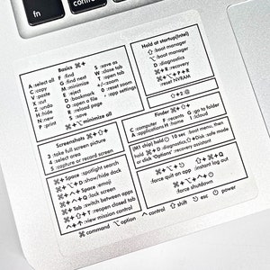 May include: A white sticker with black text detailing Mac keyboard shortcuts. The sticker is on a silver laptop. Text includes shortcuts for basics, screenshots, and system functions. The sticker is designed to help users learn and remember keyboard commands.