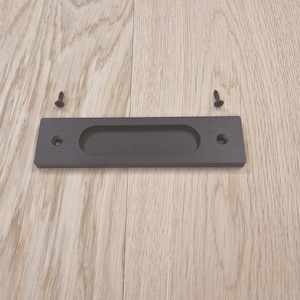 May include: A dark grey recessed door pull with an oval finger grip, and two black screws. The hardware is set against a light wood grain surface, suggesting a modern design.