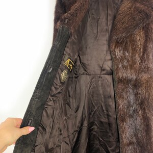 May include: A brown suede and fur coat with a brown lining. The coat has a fur collar and a button closure. There are two small metal tags on the inside of the coat.