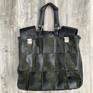May include: A black and dark olive green leather tote bag featuring a patchwork design of alternating smooth and suede textures. The bag includes two top handles, two front flap pockets with silver-toned metal push-lock closures, and a structured rectangular body.