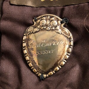 May include: Silver-toned metal tag with a heart-shaped design and a floral border. The tag has the text "God Bless" and the number "533367" engraved on it.