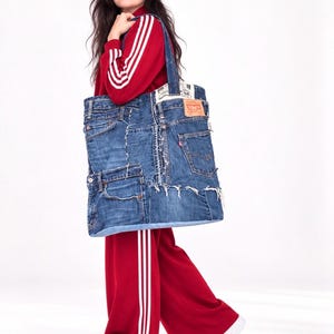 Può includere: Una grande borsa tote realizzata con jeans in denim blu riciclati, caratterizzata da tasche visibili, bordi sfrangiati e un'etichetta in pelle marrone sulla parte superiore. La borsa presenta due robusti manici in denim.