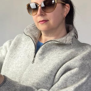 May include: A woman wearing a gray sweater with a zipper and brown cat-eye sunglasses.