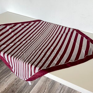May include: A burgundy and white striped scarf with a burgundy border. The scarf is folded in a triangle shape and is lying on a white surface.