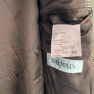 May include: A brown and black plaid blazer with a brown lining. The lining has a label with the text "BALMAIN" and the following information: "100% LAINE", "DOUBLURE: 100% VISCOSE", "5 76027", "38365/44", "TAILLE: 52", "025PBD3".