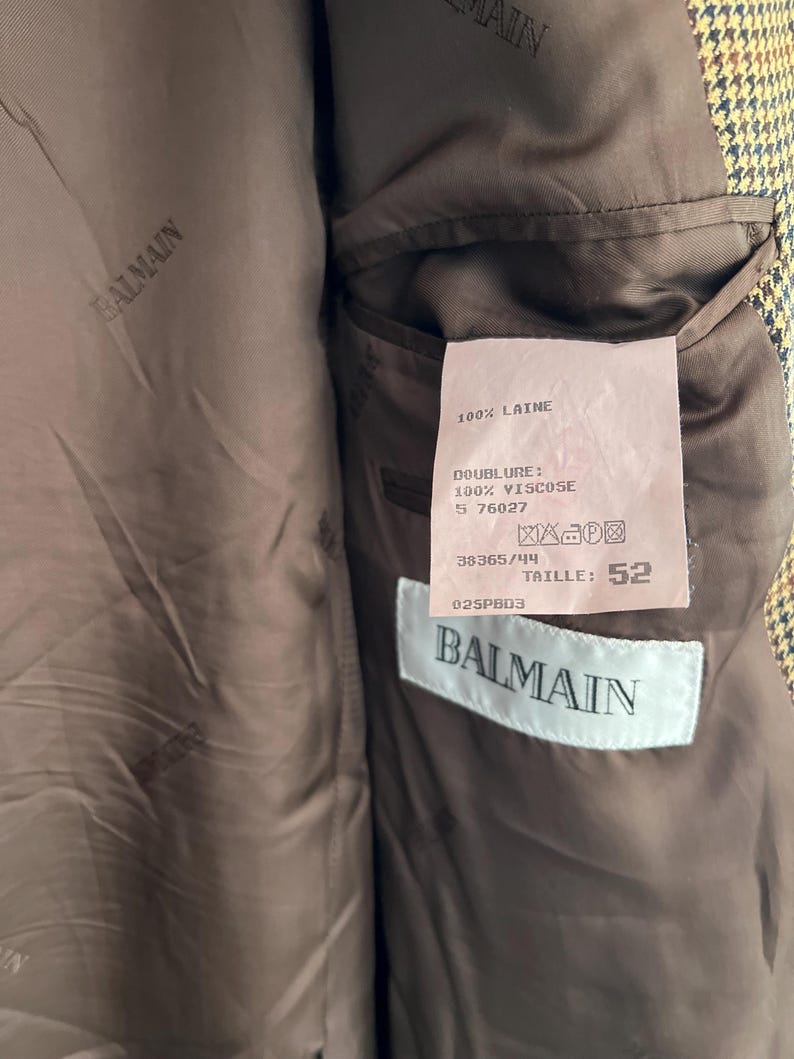 May include: A brown and black plaid blazer with a brown lining. The lining has a label with the text "BALMAIN" and the following information: "100% LAINE", "DOUBLURE: 100% VISCOSE", "5 76027", "38365/44", "TAILLE: 52", "025PBD3".