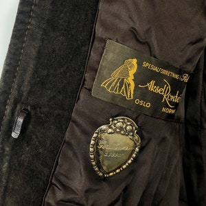May include: A brown suede jacket with a label that reads "Spesialforretning i Pels Aksel Konte Oslo NORW" and a silver heart-shaped charm with the text "Gull & S&oslash;lv 935/000".