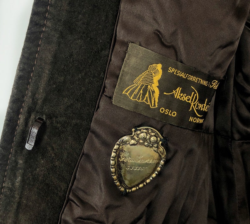 May include: A brown suede jacket with a label that reads "Spesialforretning i Pels Aksel Konte Oslo NORW" and a silver heart-shaped charm with the text "Gull & S&oslash;lv 935/000".