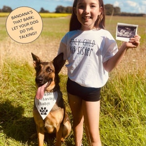 May include: A girl wearing a white t-shirt with the text "Only Child" crossed out and "Big Sister" written below it. She is holding an ultrasound image in her right hand. A German Shepherd dog is sitting next to her wearing a bandana that says "What's having a baby!"