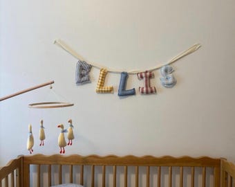May include: A fabric letter garland spelling the name ELLIS in various gingham patterns, including blue, yellow, and red, suspended on a white ribbon. Below, a wooden mobile features four hanging plush goose figures with orange feet.