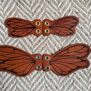 May include: Two sets of brown leather butterfly wing-shaped embellishments. Each wing has black etched details and metal eyelets for attachment. The wings are displayed on a textured, neutral-toned background.
