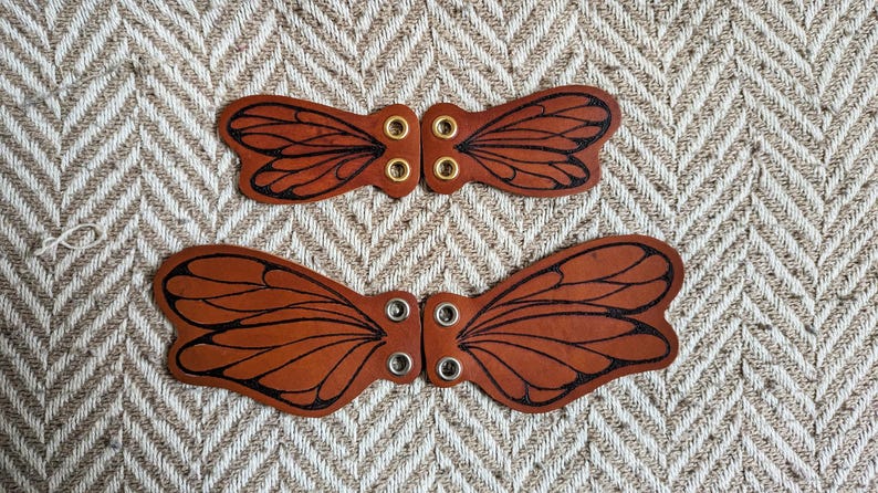 May include: Two sets of brown leather butterfly wing-shaped embellishments. Each wing has black etched details and metal eyelets for attachment. The wings are displayed on a textured, neutral-toned background.