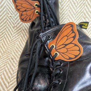 May include: Black leather boots with butterfly wing-shaped leather accents. The butterfly wings are orange with black detailing and gold-colored grommets. The boots have black laces.
