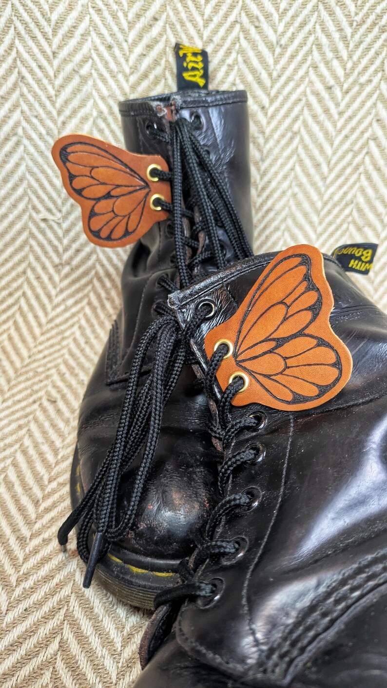 May include: Black leather boots with butterfly wing-shaped leather accents. The butterfly wings are orange with black detailing and gold-colored grommets. The boots have black laces.