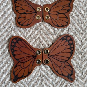 May include: Two brown leather butterfly-shaped embellishments with black etched details and brass eyelets. The wings have a detailed vein pattern. The items are displayed on a textured, neutral-toned background.