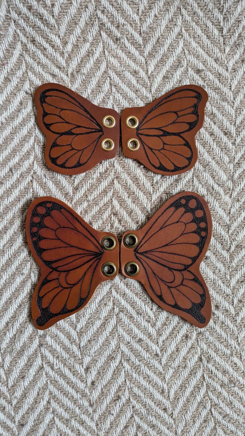 May include: Two brown leather butterfly-shaped embellishments with black etched details and brass eyelets. The wings have a detailed vein pattern. The items are displayed on a textured, neutral-toned background.