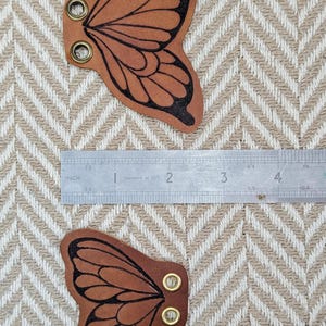 May include: Two brown leather butterfly wing shapes with black outlines and brass eyelets. A metal ruler shows measurements in inches. The wings are approximately 2 inches wide.
