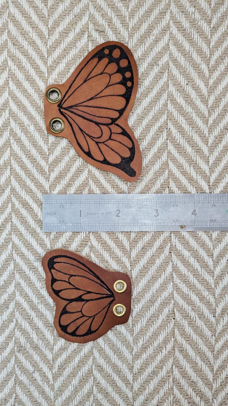 May include: Two brown leather butterfly wing shapes with black outlines and brass eyelets. A metal ruler shows measurements in inches. The wings are approximately 2 inches wide.