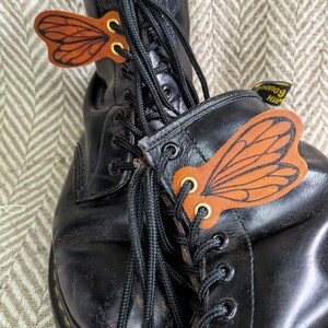 May include: Black leather boots with decorative leather butterfly wing accents. The wings are brown with orange detailing and gold eyelets. The boots have black laces and yellow stitching on the soles.