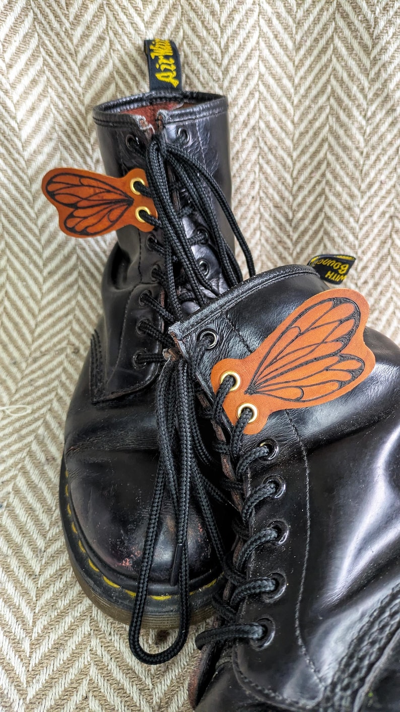 May include: Black leather boots with decorative leather butterfly wing accents. The wings are brown with orange detailing and gold eyelets. The boots have black laces and yellow stitching on the soles.