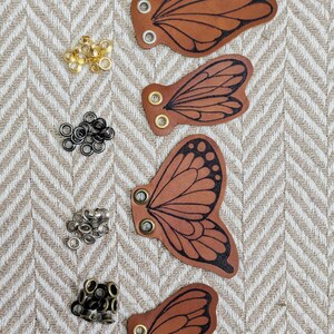 May include: Four brown leather butterfly wing cutouts with black vein detailing and metal eyelets. Various metal eyelets in gold, black, and silver are scattered around the wings. The wings are arranged on a beige and white herringbone pattern.