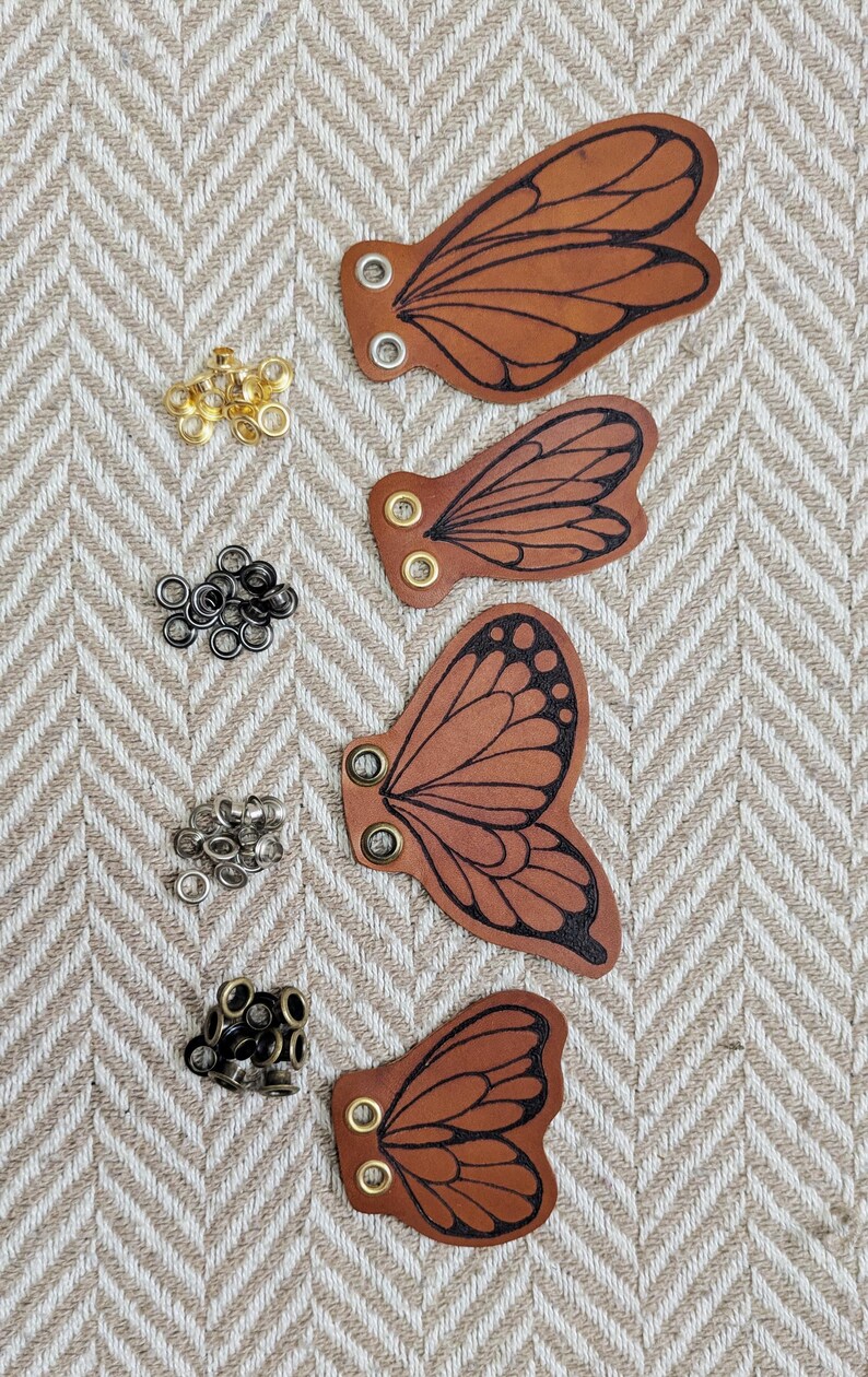 May include: Four brown leather butterfly wing cutouts with black vein detailing and metal eyelets. Various metal eyelets in gold, black, and silver are scattered around the wings. The wings are arranged on a beige and white herringbone pattern.