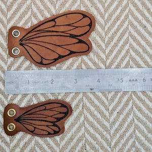 May include: Two brown leather butterfly wing-shaped patches with black wing details and metal eyelets. A metal ruler is placed next to the patches, measuring up to 9 inches. The background is a beige and white chevron pattern.