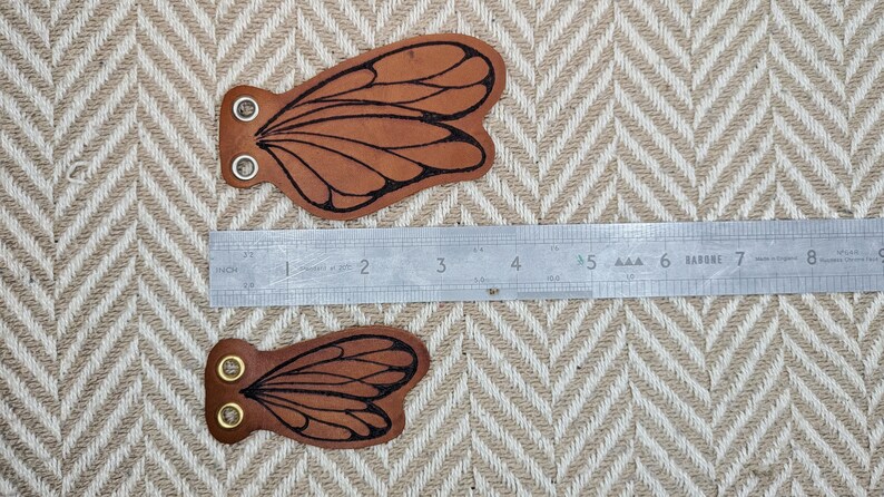 May include: Two brown leather butterfly wing-shaped patches with black wing details and metal eyelets. A metal ruler is placed next to the patches, measuring up to 9 inches. The background is a beige and white chevron pattern.